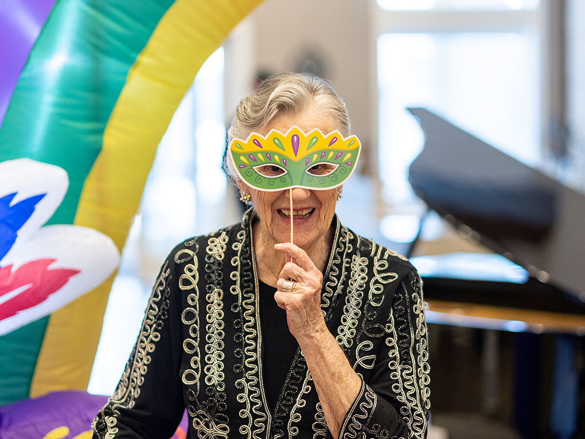 At a Mardi Gras community event, a senior resident smiles, holding up a fun mask.