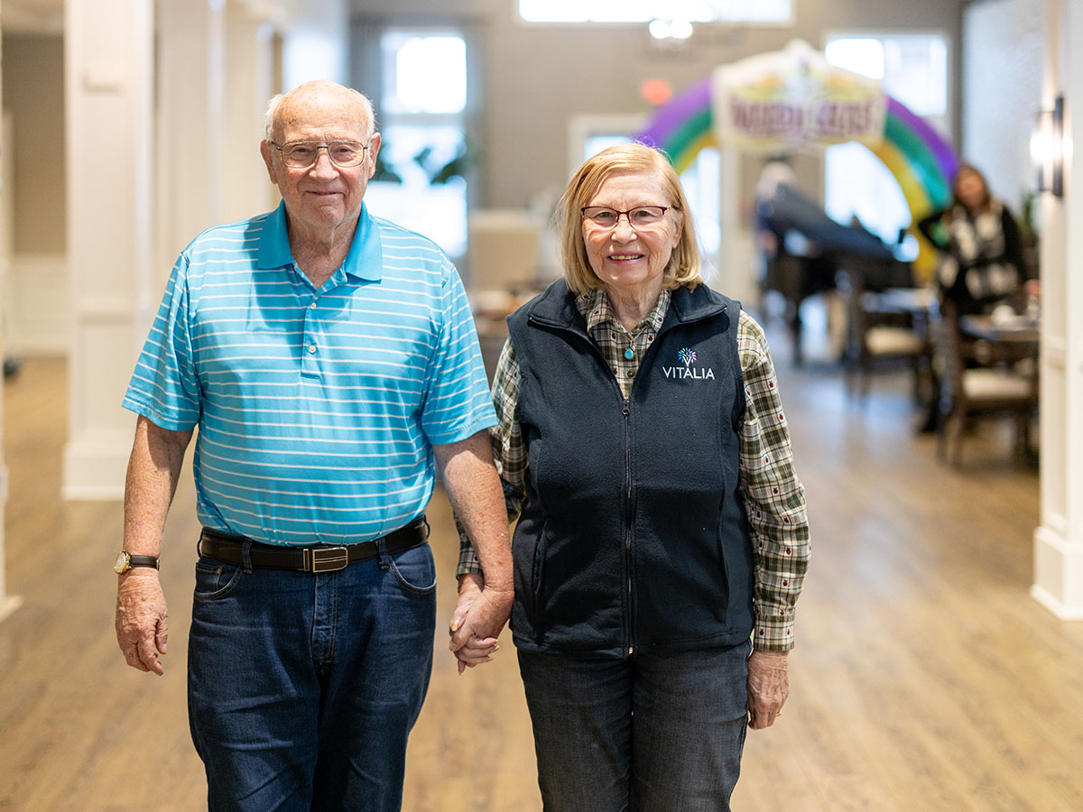 An older couple holding hands as they walk together in a hallway.