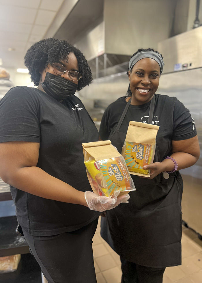 Two smiling women in black shirts and aprons hold up brown paper bags labeled 'You Rock!' with colorful snacks inside, standing in a kitchen environment.