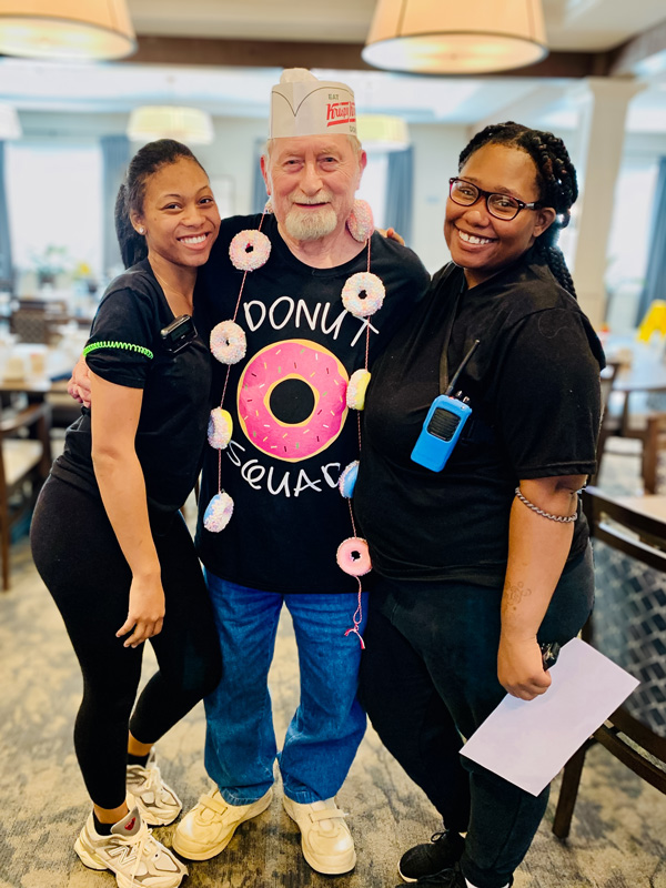 A senior resident dressed in a 'Donut Squad' shirt and Krispy Kreme hat poses with two smiling staff members during a fun-themed event at a senior living community.