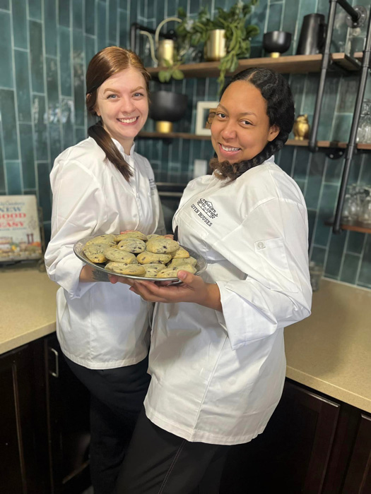 Two smiling chefs in white uniforms proudly present a tray of freshly baked cookies in a modern bistro kitchen with green tile walls, ready to serve at a senior living community.