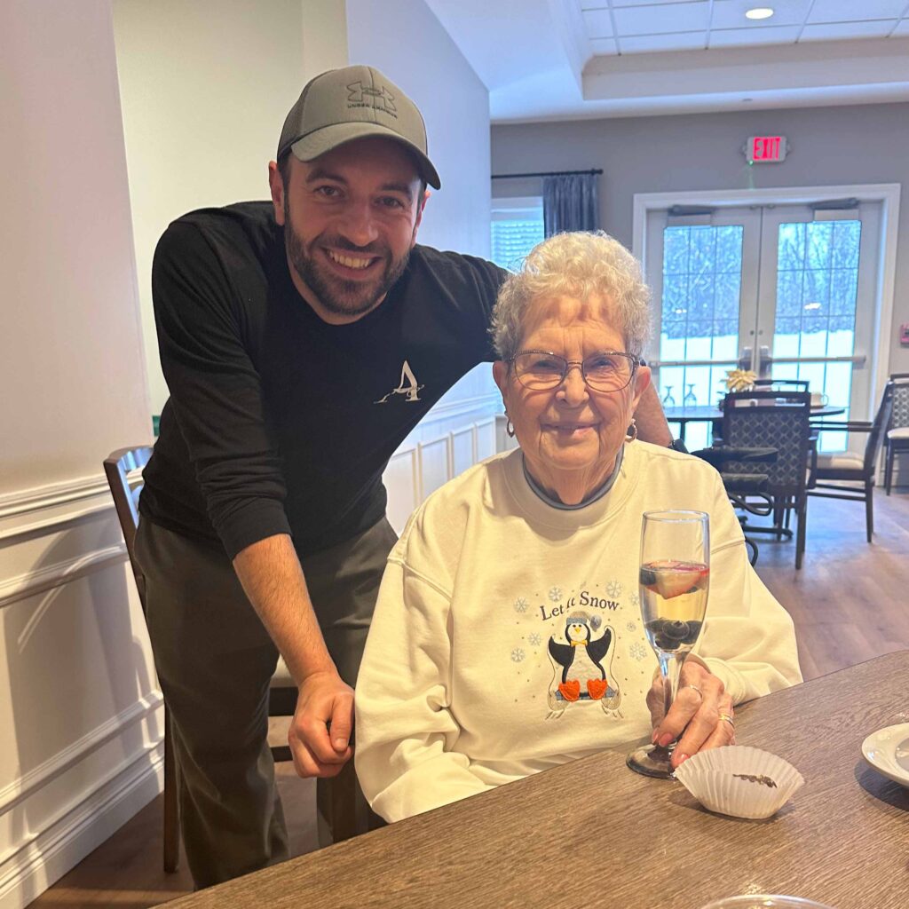 A Culinary Director poses with a smiling resident wearing a festive sweater, holding a glass of sparkling drink during a winter celebration in the dining area.