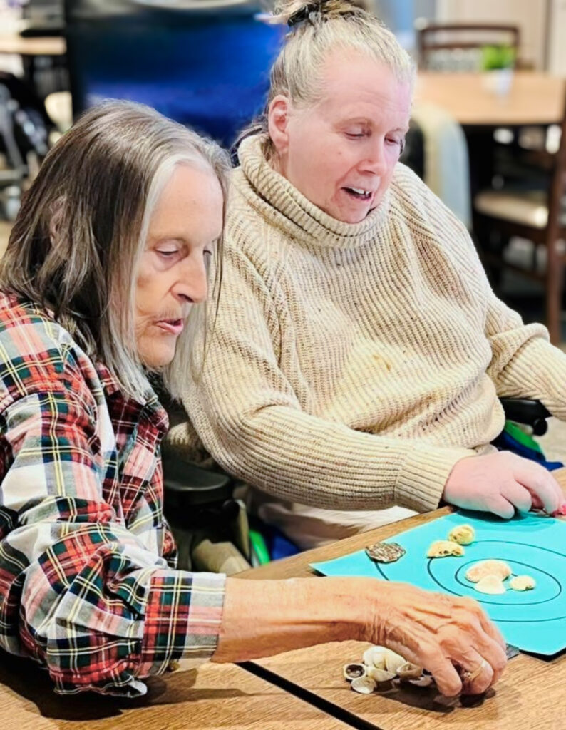 Two women enjoy a hands-on activity together at a senior living community, arranging seashells on a blue board while sharing focus and companionship.