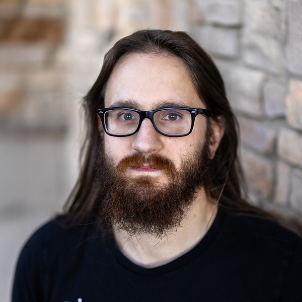 Jesse Hollis, Plant Operations Director at Vitalia Montrose, standing beside a stone wall. He has long brown hair, a full beard, and glasses, and is wearing a black shirt in this professional portrait.