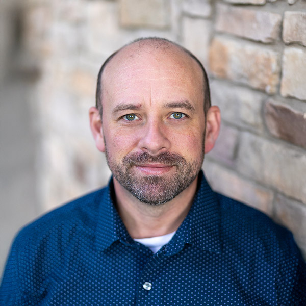 Robert Miller, Operations Coordinator at Vitalia Montrose, smiling in a professional headshot while standing beside a stone wall. He is wearing a navy patterned shirt layered over a white undershirt.