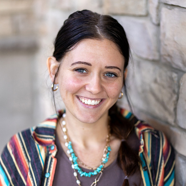 Whitney Wilson, Memory Care Director at Vitalia Montrose, smiling warmly while standing against a stone wall. She wears a striped colorful cardigan over a brown top with turquoise jewelry and has her hair styled in a side braid.