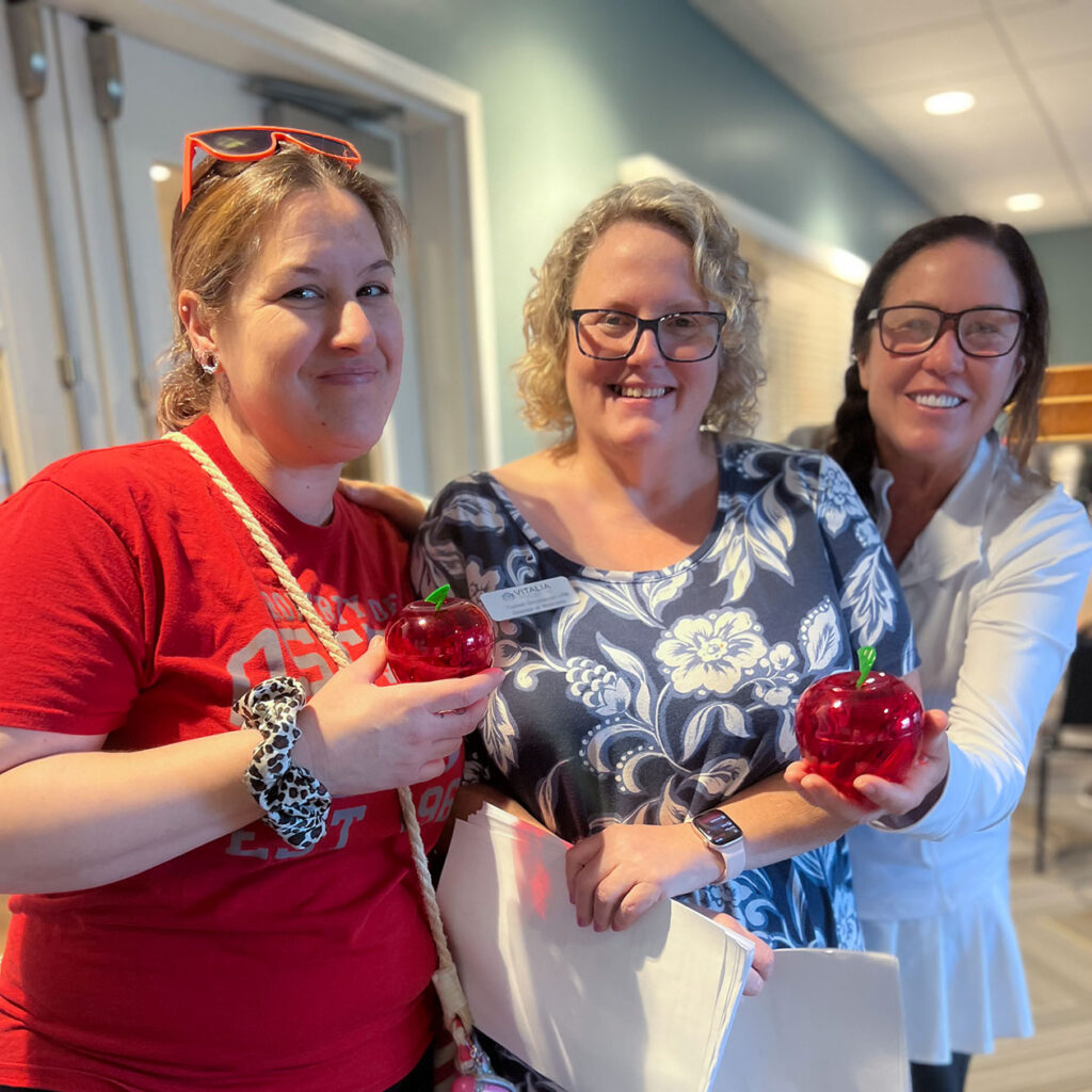 Three women smile brightly, two holding novelty apple gifts during an event.