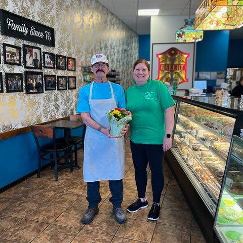 Owners at Tiffany's Bakery smile proudly, holding a gift of potted flowers from Vitalia Montrose.