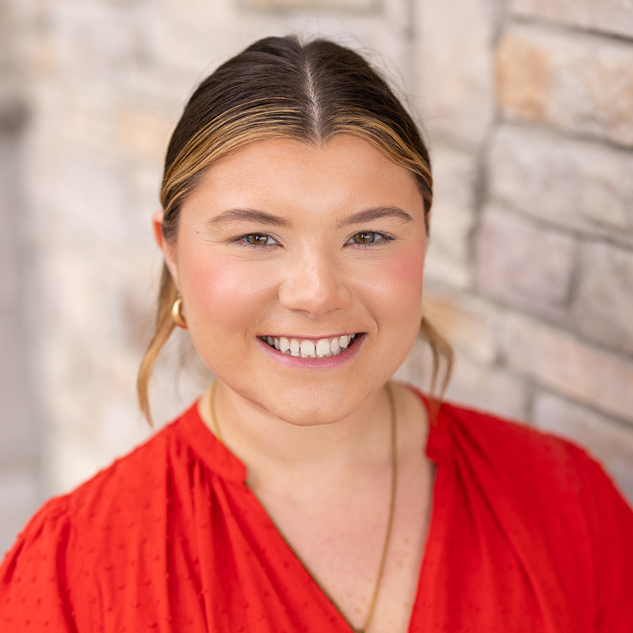 Jessica Standly, Villa Leasing Coordinator for Vitalia Montrose and The V Living, smiling in a professional headshot with highlighted hair pulled back, a red blouse, gold hoop earrings, and a softly blurred light stone background.