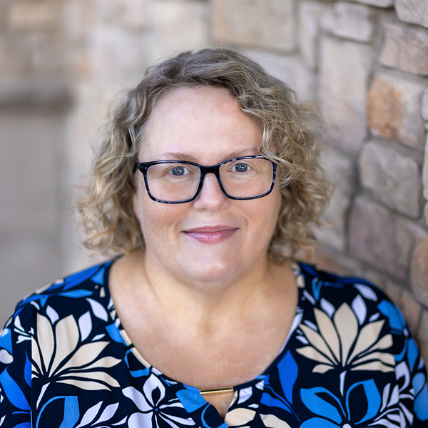 Tommi Smallwood, Wellness Director at Vitalia Montrose, smiling in a professional portrait beside a stone wall. She has curly blonde hair, wears glasses, and is dressed in a blue and cream floral blouse.