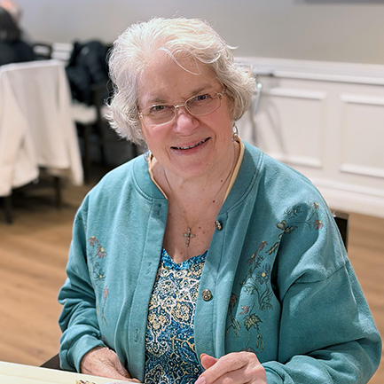 A senior woman wearing a blue cardigan smiles welcomingly at Vitalia Montrose.