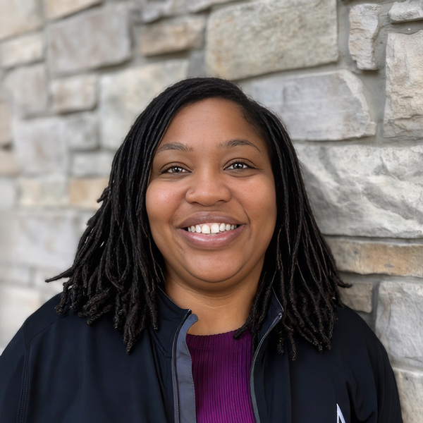 Akita Orr, Memory Care Director at Vitalia Montrose, smiling in a professional headshot with shoulder-length locs, a magenta sweater, a black jacket, and a stacked stone wall background