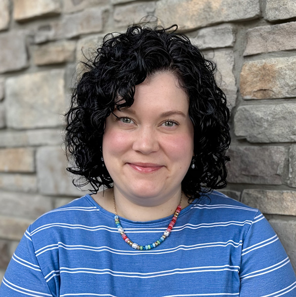 Calie King, Operations Coordinator at Vitalia Montrose, looking at the camera in a professional headshot with short curly dark hair, a blue striped top, a colorful beaded necklace, and a stacked stone wall background.