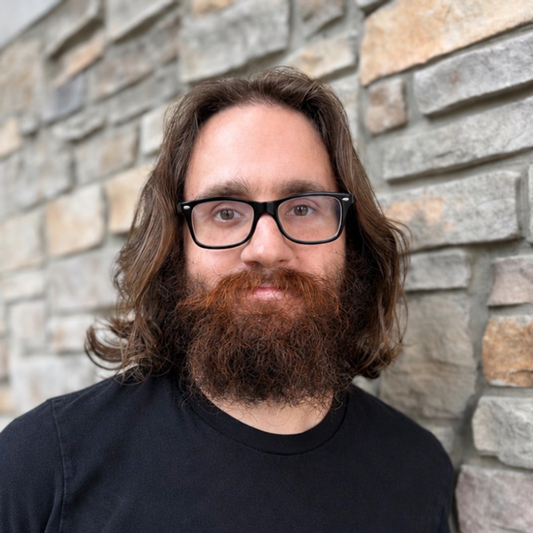 Jesse Hollis, Plant Operations Director at Vitalia Montrose, looking at the camera in a professional headshot with long wavy brown hair, glasses, a full beard, a black shirt, and a stacked stone wall background.